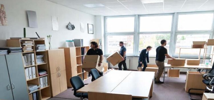 Modern irodai tér költöztetés közben: munkatársak dobozokat pakolnak, irodabútorok és kartondobozok szétszórva a helyiségben nagy ablakokkal és természetes fénnyel.